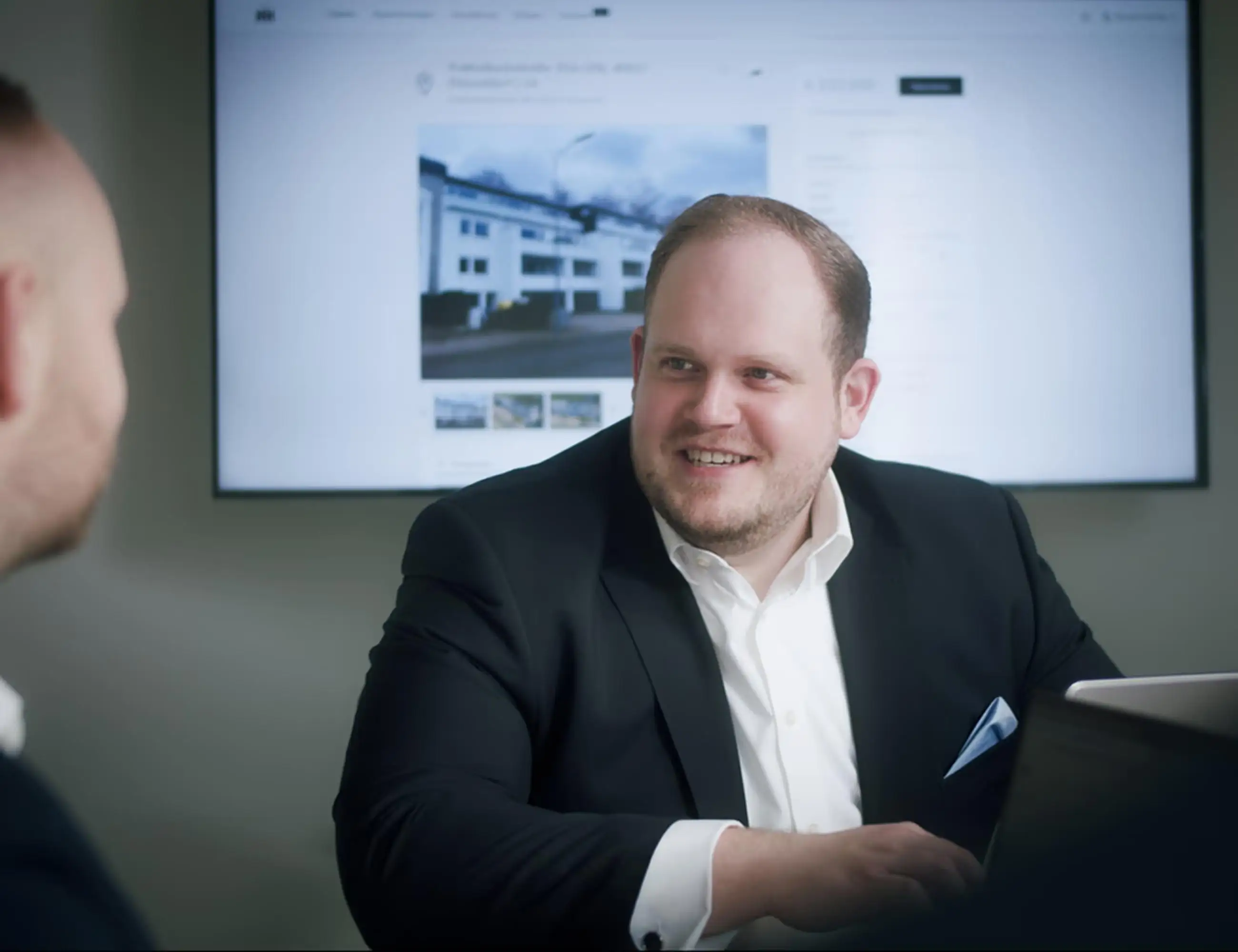 Ein Mann in Anzug und weißem Hemd sitzt lächelnd vor einem Laptop, im Hintergrund ein Immobilienfoto.
